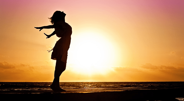 Aplicativo para fortalecer a autoestima. Na foto, uma mulher de vestido está feliz, com os braços abertos, com o pôr do sol ao fundo