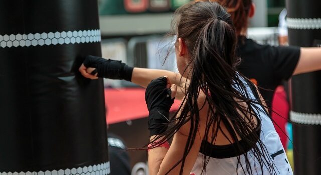 Para combater o estresse, é muito importante fazer exercícios físicos. Na foto, uma mulher está em uma academia, com o cabelo preso e suado, usando roupas de ginástica, dando socos em um saco de pancadas